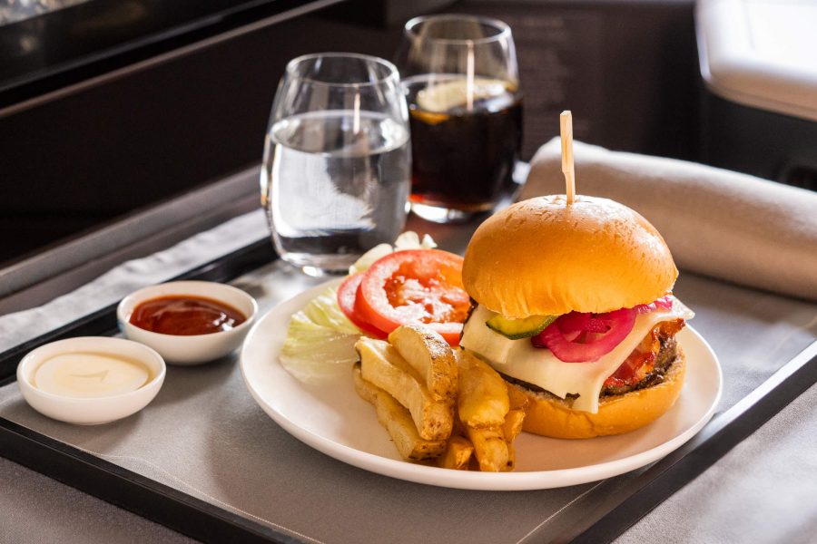 A classic beef burger is served on a white plate with thick-cut fries, tomato slices and lettuce, accompanied by two small condiment dishes and beverages. The meal is presented on a tray table inside a Cathay Pacific aircraft. 
